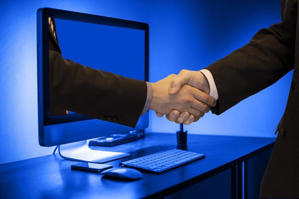 デスクトップPCの画面から出てきた手と握手するビジネスパーソン